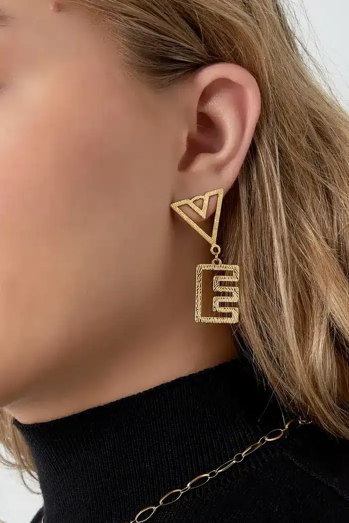 Nahaufnahme des Ohrs einer Person, die mit exquisiten Love-Ohrsteckern geschmückt ist. Das geometrische Ohrhänger-Design weist eine dreieckige Form auf, die mit einer rechteckigen Form mit aufwendigen Aussparungsdetails verbunden ist. Die Person mit hellbraunem Haar und einem schwarzen Rollkragenpullover strahlt eine Aura der Eleganz und eine Liebe für schicke Accessoires aus.