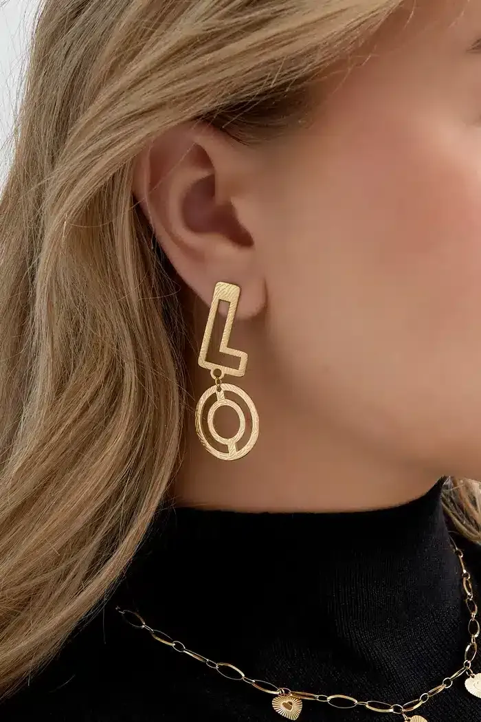 Nahaufnahme einer Person, die den Love-Ohrstecker trägt, einen goldenen Ohrring in Form des Buchstabens „L“, der über einem kreisförmigen Design hängt. Der Kopf der Person ist leicht zur Seite gedreht und zeigt blondes Haar und einen Teil eines schwarzen Rollkragenpullovers. Eine goldene Halskette verleiht einen zusätzlichen Hauch von Liebe und ergänzt den eleganten Love-Ohrstecker perfekt.