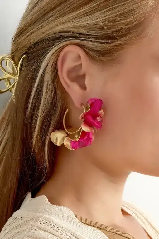 Nahaufnahme des Ohrs einer Person, das mit großen, dekorativen Herbstblumen-Ohrsteckern in Gold- und leuchtenden Rosatönen geschmückt ist. Die Person hat langes, blondes Haar, das mit einer goldenen Haarspange befestigt ist, und der Saum eines beigen Kleidungsstücks ist ebenfalls sichtbar.