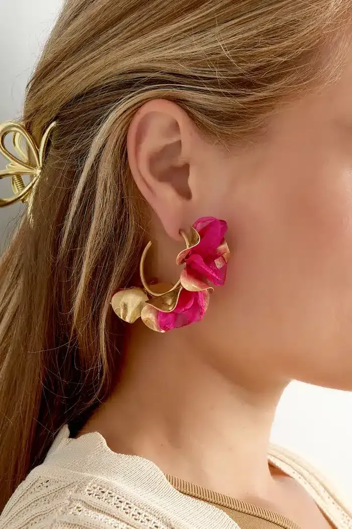 Nahaufnahme des Ohrs einer Person, das mit großen, dekorativen Herbstblumen-Ohrsteckern in Gold- und leuchtenden Rosatönen geschmückt ist. Die Person hat langes, blondes Haar, das mit einer goldenen Haarspange befestigt ist, und der Saum eines beigen Kleidungsstücks ist ebenfalls sichtbar.