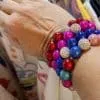 Ein mit mehreren Magic Beads Ina Armband-Armbändern geschmücktes Handgelenk, das bunte runde Perlen in Rot-, Blau-, Rosa- und Lilatönen zeigt, die mit funkelnden Silberperlen durchsetzt sind. Am Ringfinger ist ein goldener Ring zu sehen, während im Hintergrund Kleidungsstücke hängen.