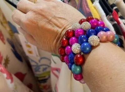 Ein mit mehreren Magic Beads Ina Armband-Armbändern geschmücktes Handgelenk, das bunte runde Perlen in Rot-, Blau-, Rosa- und Lilatönen zeigt, die mit funkelnden Silberperlen durchsetzt sind. Am Ringfinger ist ein goldener Ring zu sehen, während im Hintergrund Kleidungsstücke hängen.