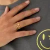 Eine Person, die ein graues Hemd mit einem gelben Smiley-Design trägt, zeigt ihre Hand, an der ein Oh Happy Day-Ring hängt – ein goldener Ring mit zwei Kreisen, die jeweils einen Buchstaben „G“ enthalten. Der goldene Ring steckt am Mittelfinger der Hand.