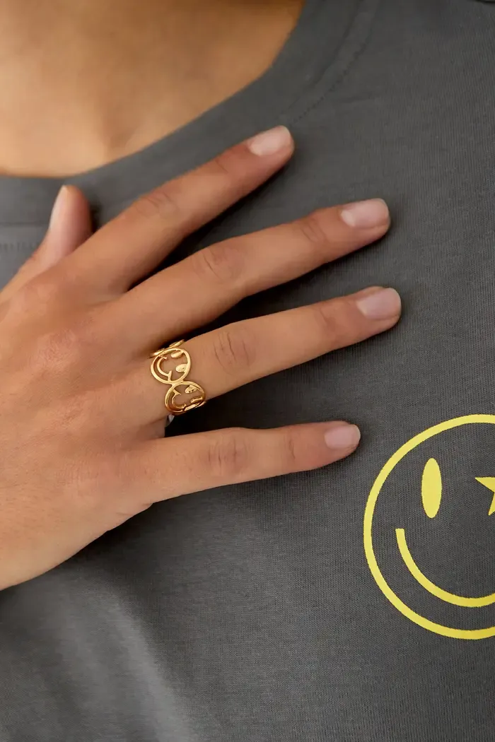 Eine Person, die ein graues Hemd mit einem gelben Smiley-Design trägt, zeigt ihre Hand, an der ein Oh Happy Day-Ring hängt – ein goldener Ring mit zwei Kreisen, die jeweils einen Buchstaben „G“ enthalten. Der goldene Ring steckt am Mittelfinger der Hand.