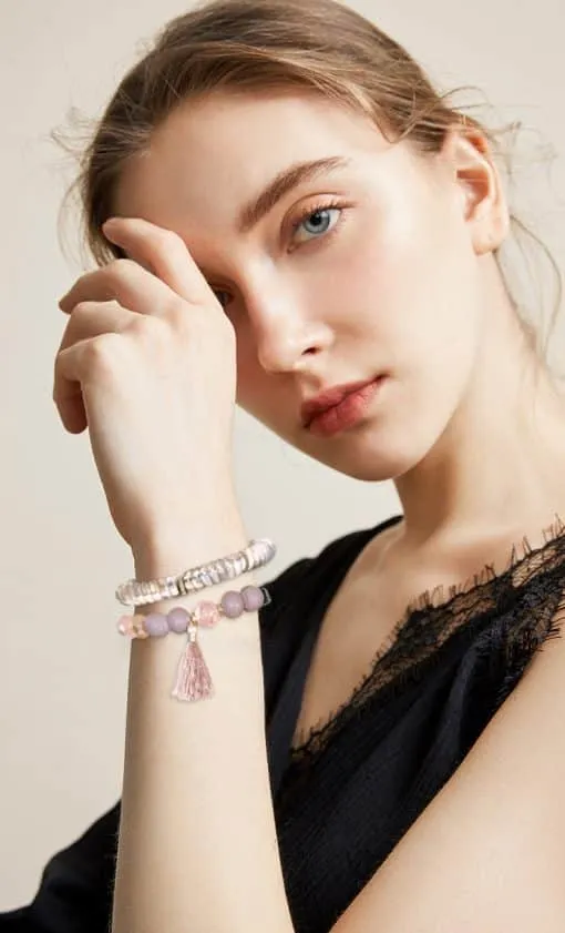 Eine Person mit heller Haut und hellbraunem Haar hält ihre Hand vor ihr Gesicht und präsentiert das **Buddha Love Armband**. Das Armband ist mit rosa und weißen Perlen und einer rosa Quaste verziert und strahlt Liebe aus. Die Person trägt ein schwarzes, strukturiertes ärmelloses Oberteil.