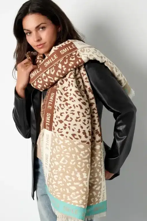 Eine Person mit langen dunklen Haaren trägt eine schwarze Lederjacke und einen Wild Smile Schal (Kopie) mit Leopardenmuster und „SMILE“ in leuchtendem Türkis. Sie steht selbstbewusst vor einem schlichten weißen Hintergrund.