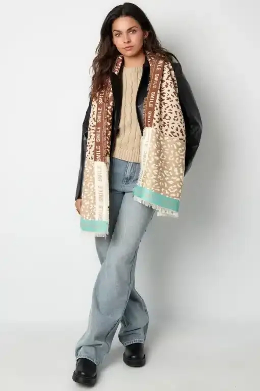 Eine Person mit langen dunklen Haaren steht vor einem schlichten Hintergrund und trägt eine Lederjacke, einen beigen Pullover, blaue Jeans und schwarze Schuhe. Über ihren Schultern hängt der Fashion Smile Schal mit braunen und hellblauen Akzenten, die ihr stilvolles Modestatement unterstreichen, während sie sanft lächelt.