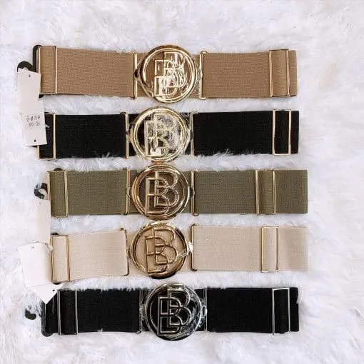 Es werden fünf BB Stretch Gürtel ausgestellt, die große runde Metallschnallen mit einem überlappenden Buchstabendesign aufweisen. Die Gürtel sind in Beige, Schwarz, Grün und Taupe erhältlich und ordentlich auf einer strukturierten weißen Oberfläche angeordnet.
