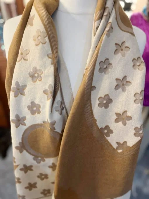 Eine Schaufensterpuppe präsentiert den „Cherrys“-Schal, einen beige-braunen Schal, der mit floralen und abstrakten Mustern verziert ist, die an einen Kirschblütentag erinnern. Der Schal ist lässig um den Hals drapiert, was die weichen und leichten Eigenschaften des Stoffes hervorhebt.