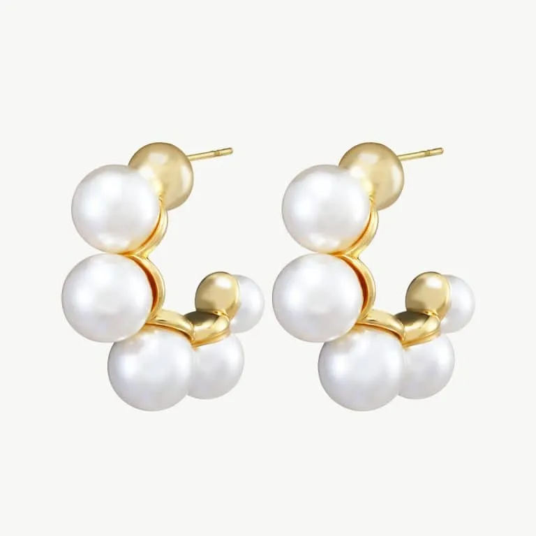 Die Perlencreolen-Ohrringe bestehen aus runden Ohrsteckern aus Gold, die mit Gruppen weißer Perlen verziert sind, die anmutig in einer Kurve angeordnet sind und einen stilvollen und eleganten Kontrast zu einem schlichten, hellen Hintergrund bilden.