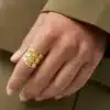 Eine Hand mit einem goldenen Dreibandring ruht auf einer Oberfläche. Die Person trägt ein grünes langärmeliges Kleidungsstück. Der Hintergrund ist verschwommen und passt farblich zur Kleidung.