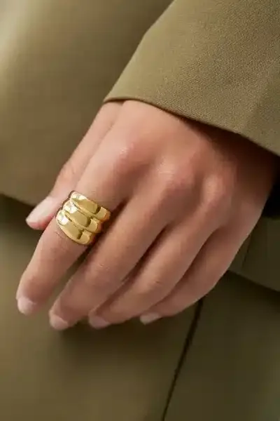 Eine Hand mit einem goldenen Dreibandring ruht auf einer Oberfläche. Die Person trägt ein grünes langärmeliges Kleidungsstück. Der Hintergrund ist verschwommen und passt farblich zur Kleidung.