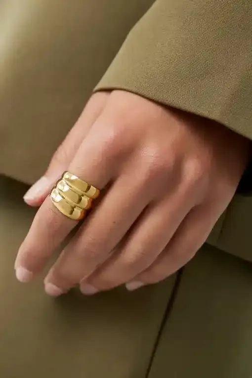 Eine Hand mit einem goldenen Dreibandring ruht auf einer Oberfläche. Die Person trägt ein grünes langärmeliges Kleidungsstück. Der Hintergrund ist verschwommen und passt farblich zur Kleidung.