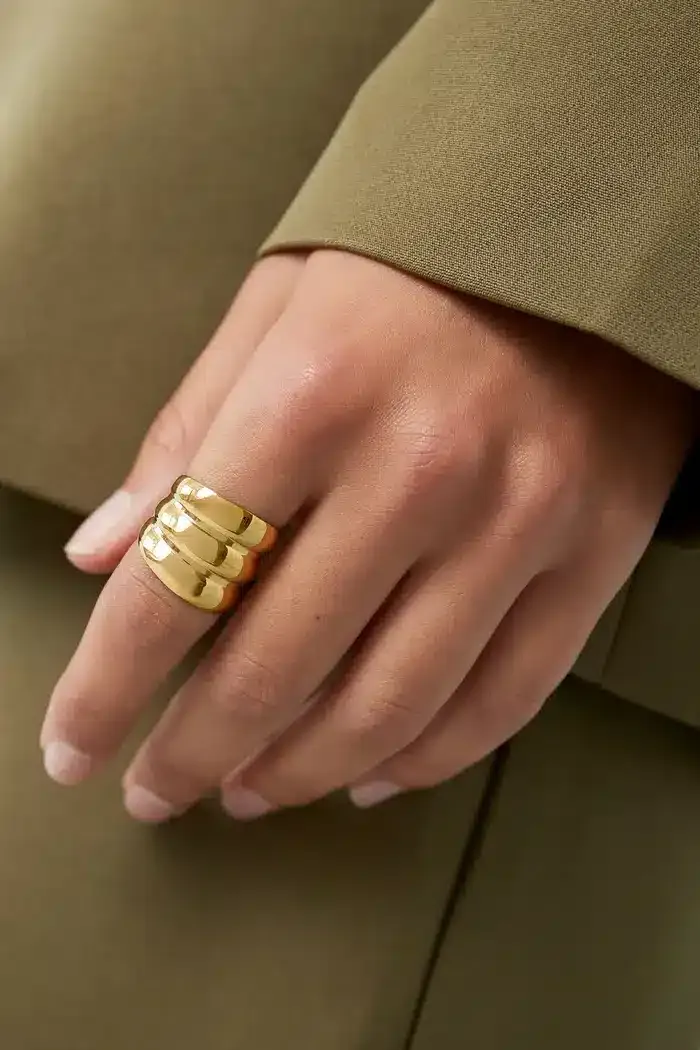 Eine Hand mit einem goldenen Dreibandring ruht auf einer Oberfläche. Die Person trägt ein grünes langärmeliges Kleidungsstück. Der Hintergrund ist verschwommen und passt farblich zur Kleidung.