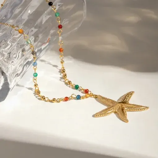 Eine goldene Halskette mit einem Seesternanhänger auf einer weißen Oberfläche. Die Kette ist mit kleinen, bunten Perlen besetzt. Man sieht Schatten, die von der Halskette und einem Glasobjekt geworfen werden.