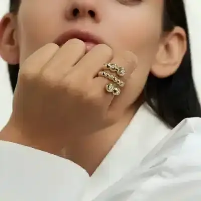 Das Bild zeigt eine Person mit langen dunklen Haaren in einem weißen Hemd, das teilweise von der unteren Gesichtshälfte aus sichtbar ist. Ihre Hand befindet sich in der Nähe ihres Mundes und zeigt zwei Spiral-Goldringe mit kreisförmigen Mustern und kleinen Edelsteinen an ihren Fingern.