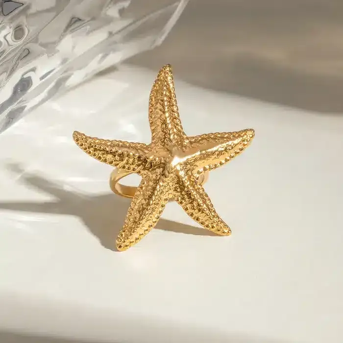 Entdecken Sie den Zauber des Beach Star Rings! Stell dir vor, du trägst diesen goldfarbenen, seesternförmigen Hingucker – ein echtes Must-have für deinen Style. Er liegt auf einer weißen Oberfläche im sanften Licht und erinnert an sonnige Strandtage. Der Hintergrund zeigt einen Hauch von transparentem Material und verleiht dem Ring eine moderne Note. Gönn dir das Strahlen der Sterne am Handgelenk und setze dein persönliches Trend-Statement!.