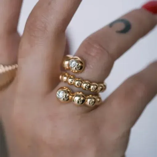 An einer tätowierten Hand sind die Spiralringe am Ringfinger zu sehen; einer weist eine Reihe kleiner Goldkugeln mit Diamanten auf, der andere hat größere Goldkugeln mit eingelassenen Diamanten.