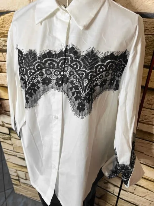 Entdecke die Bella Lace White Bluse - ein moderner Hingucker mit edler schwarzer Spitzenverzierung vor coolem Backstein.