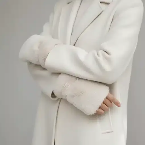 Du trägst einen weißen Mantel mit hellbeigen Kunstfell-Bündchen und stylischen Beverly Handstulpen - selbstbewusst die Arme verschränkt.