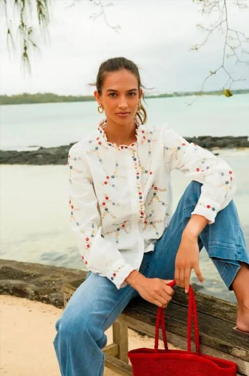 Erlebe den Sommerlook: Du sitzt entspannt am Wasser, trägst eine weiße Blumenbluse, Jeans und stylische rote Tasche.