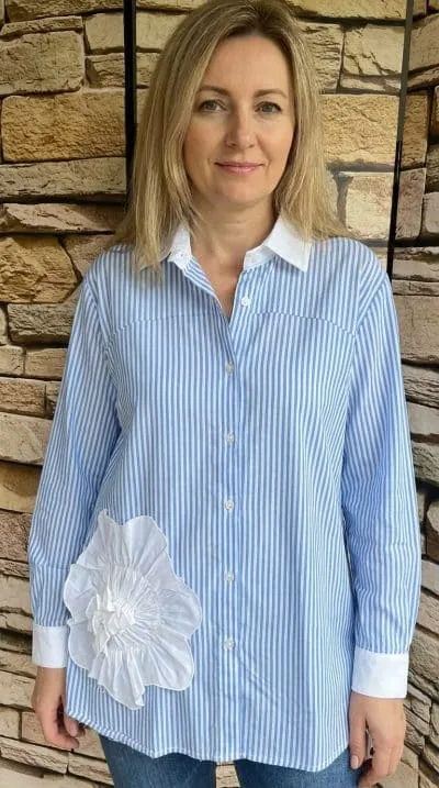 Du stehst entspannt an einer Steinmauer, trägst ein modernes blau-weiß gestreiftes Shirt mit auffälliger Blütenapplikation.