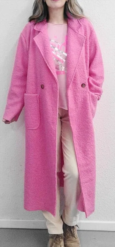 Du trägst einen langen pinken Mantel, helle Hosen und braune Schuhe - modern gestylt vor einer hellen Wand.