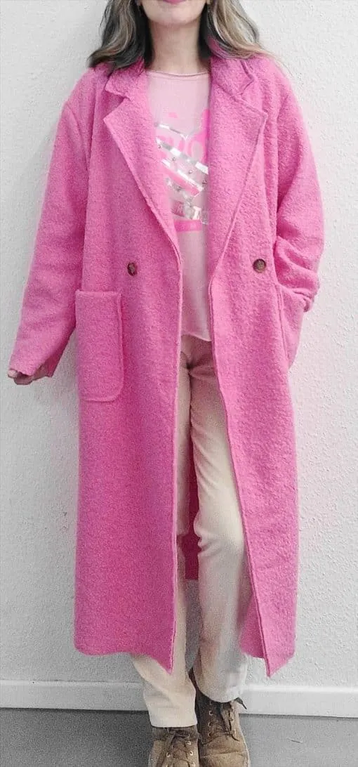 Du trägst einen langen pinken Mantel, helle Hosen und braune Schuhe - modern gestylt vor einer hellen Wand.