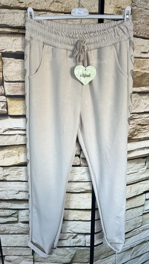 Entdecke die Heart Side Jogpants in Beige - elastischer Bund, praktische Taschen und stylisher Herz-Tag vor trendiger Steinwand.
