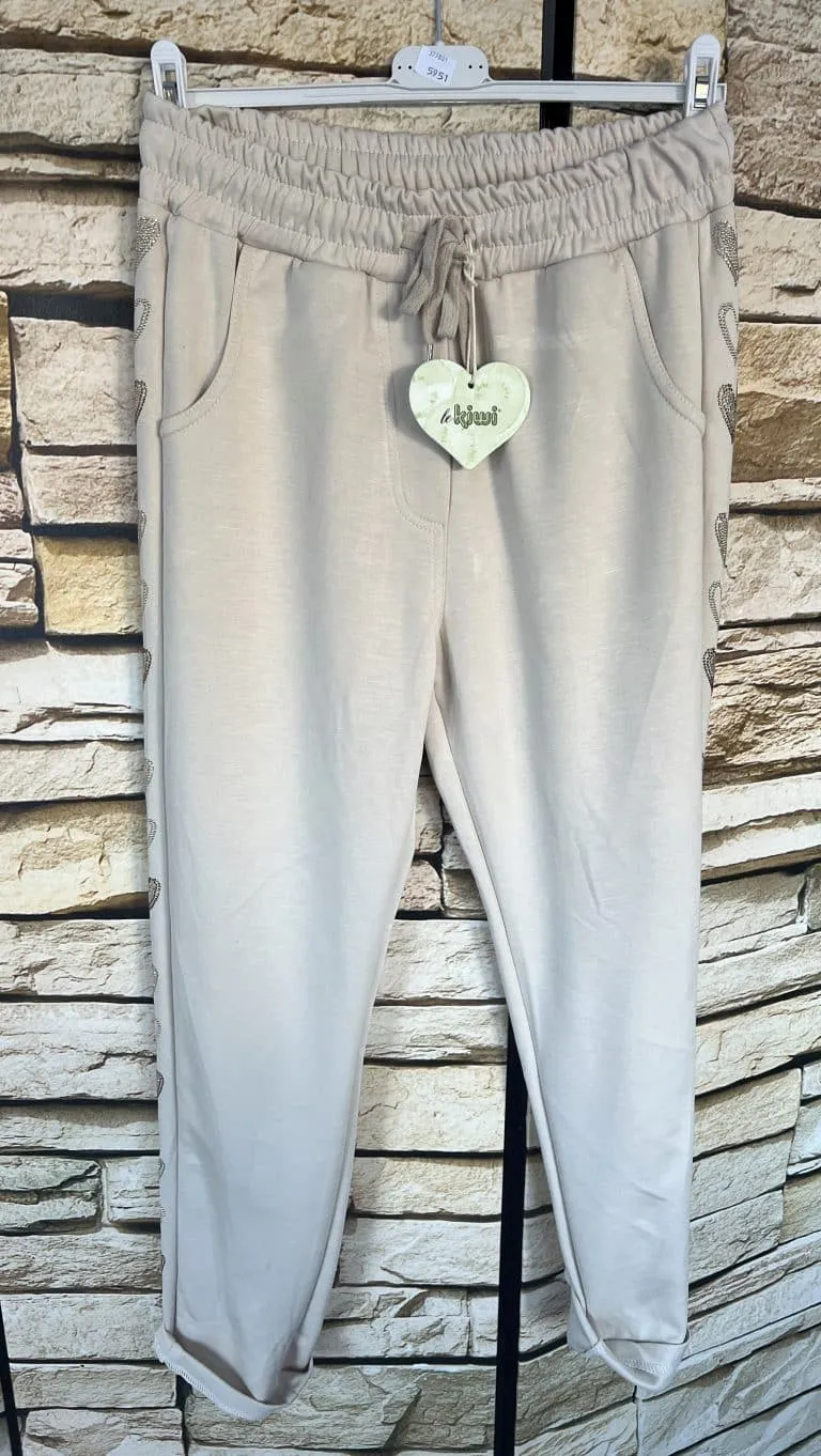Entdecke die Heart Side Jogpants in Beige - elastischer Bund, praktische Taschen und stylisher Herz-Tag vor trendiger Steinwand.
