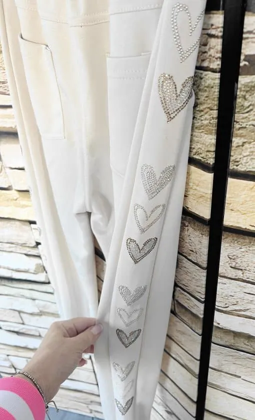 Hol dir den modernen Look: Weiße Heart Side Jogpants mit funkelnden Herz-Details, cool in deiner Hand vorm Steinwand-Hintergrund.
