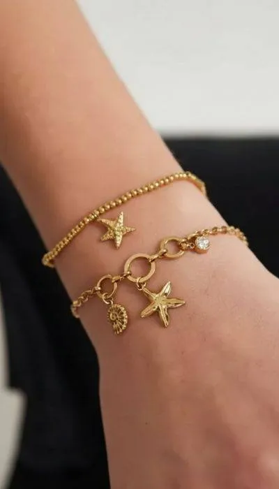Home Beach Premium Line Armband: Goldene Armbänder mit Seestern- und Muschel-Anhängern am Handgelenk.