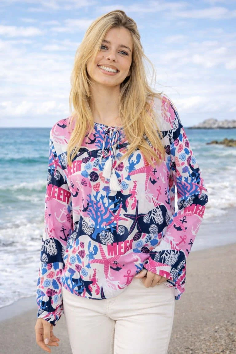 Frau am Strand in farbenfroher Maritime Mood Tunikabluse und weißer Hose - lebendiger, stilvoller Komfort, perfekt für Tage am Meer.