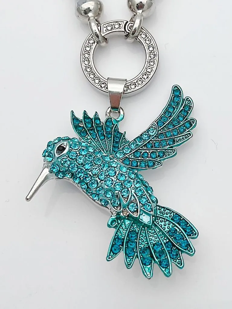 Kolibri Wechsel Anhänger: A close-up shows a hummingbird pendant with blue rhinestones on a silver chain, shimmering brightly.