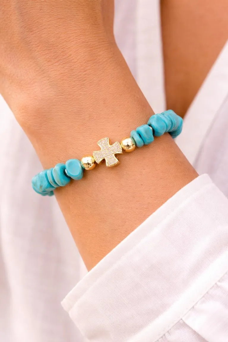 Close-up of a wrist wearing a turquoise stone bracelet with gold beads and a sparkling gold cross charm.